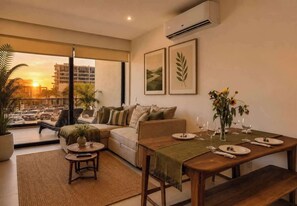 A modern living room with a sofa, a dining table set for two, and a view of a cityscape through large windows.