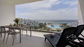 Ein Balkon mit Esstisch, einer Liege und Blick auf ein Resort mit Pool und Strand.