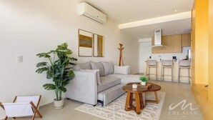 Ein modernes Wohnzimmer mit einem grauen Sofa, einem Couchtisch und einer großen Küstpflanze. Im Hintergrund ist eine Küchenzeile mit einer Bar und Hockern zu sehen.