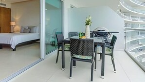 A modern hotel room with a bed, a dining table with chairs, and a glass wall leading to a balcony.