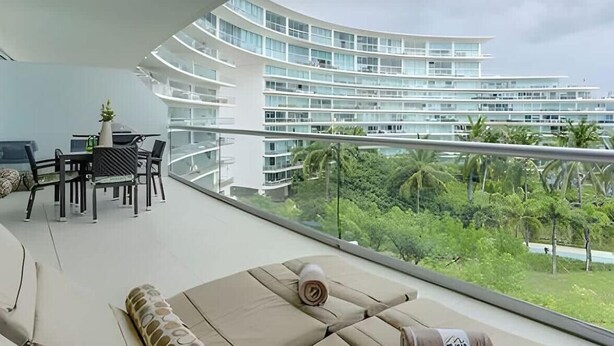 A modern hotel room with a curved balcony, a dining area with a table and chairs, and a view of a multi-story building and lush greenery.