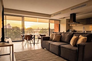 A modern living room with a large window, a sofa, a dining table with chairs, and a kitchen area.