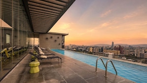 Ein Dachpool mit Liegestühlen und Fitnessgeräten mit Blick auf eine Stadtlandschaft bei Sonnenuntergang.