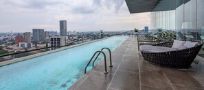 A rooftop infinity pool with a city view, a glass railing, and a wicker lounge chair.