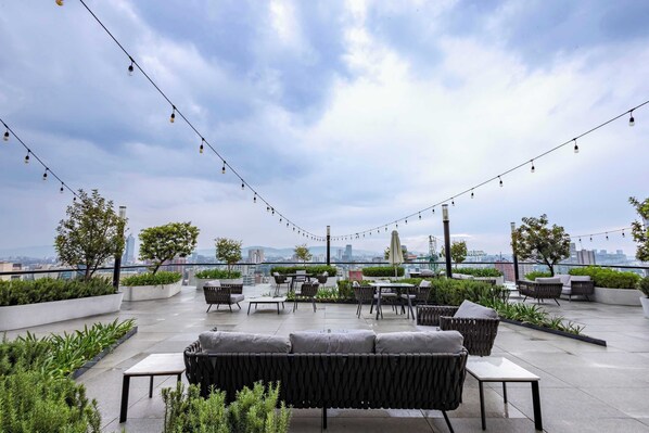 Eine Dachterrasse mit Sitzgelegenheiten im Freien, Lichterketten und Blick auf die Stadt.