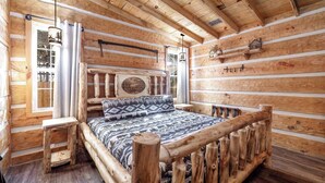 Een slaapkamer in een blokhut met een houten bed, een klein tafeltje en een raam met gordijnen.