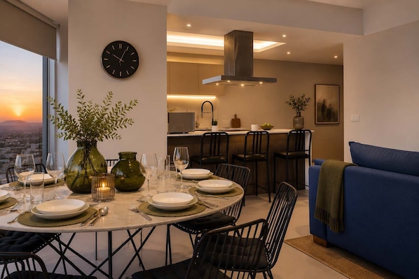 A dining area with a table set for four, a clock on the wall, and a city view through the window.