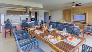 Una sala de estar moderna con una mesa de comedor de madera dispuesta para el desayuno, mobiliario de mimbre azul, un televisor grande y una zona de cocina al fondo.