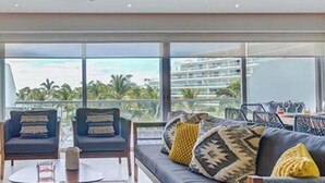 Una sala de estar moderna con una televisión de pantalla plana, dos sillones, un sofá y un balcón con vistas a palmeras y edificios.