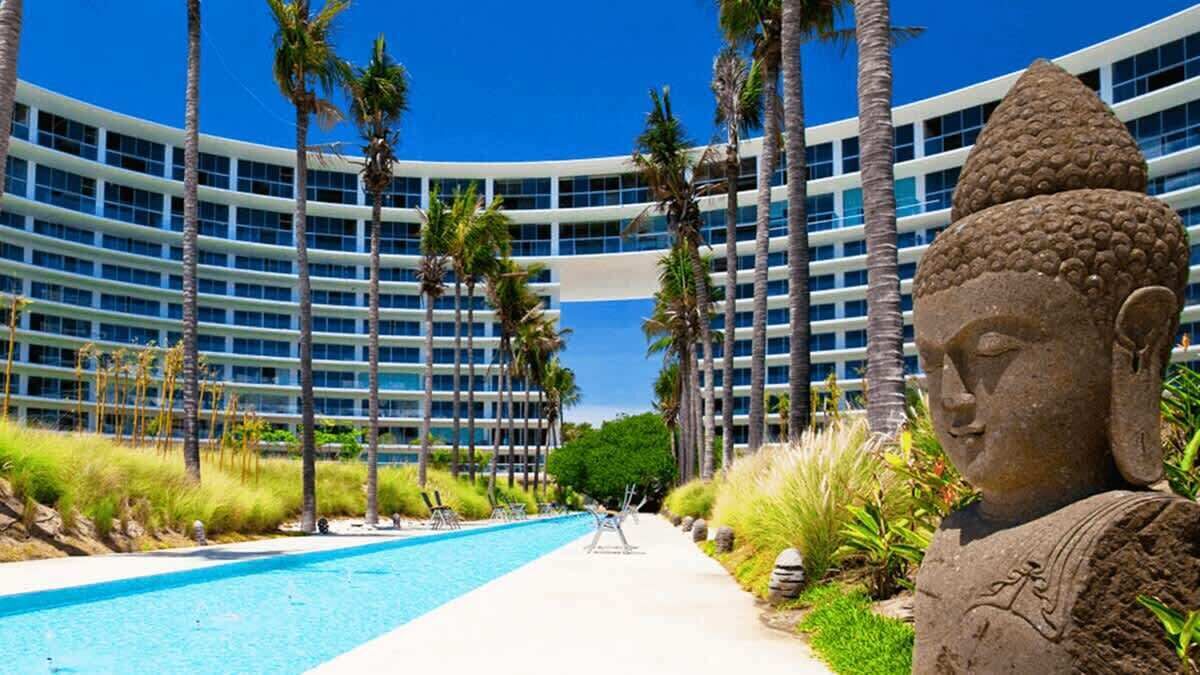 A modern hotel with a pool, palm trees, and a Buddha statue.