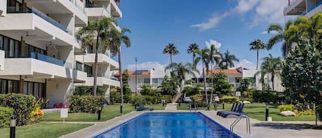 A modern hotel with a swimming pool, palm trees, and a clear blue sky.