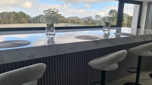 Uma área de bar moderna com bancada de mármore, dois vasos de vidro com flores brancas e vista para uma paisagem natural através de grandes janelas.