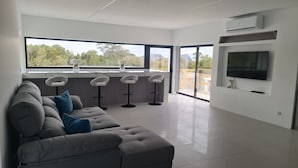 Uma sala de estar moderna com um sofá cinzento, um espaço de cozinha com balcão e uma TV fixada na parede.