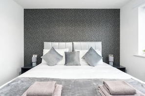 A modern hotel room with a large bed, grey textured wall, and two bedside tables with lamps. - Stunning & Spacious Home | Driveway | Sleeps 6! (Sleaford)