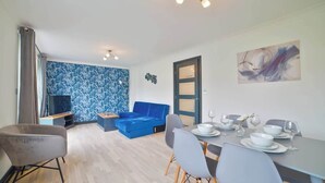 Un salon moderne avec un canapé bleu, une table à manger dressée pour quatre personnes et une grande fenêtre avec des rideaux.
