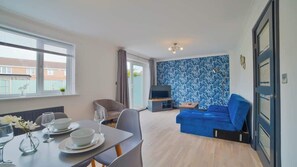 A modern living room with a dining area, a blue sofa, a TV, and a patterned wall. - Stunning & Spacious Home | Driveway | Sleeps 6! (Sleaford)