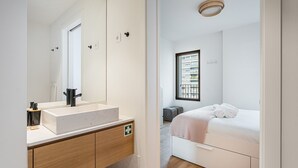 Ein modernes Badezimmer mit einem großen Spiegel, einem weißen Waschbecken und einem Holzkabinet. Ein Fenster bietet Blick auf Gebäude.