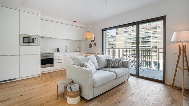 Eine moderne Wohnung mit einer Küchenzeile, einem grauen Sofa, einem kleinen runden Hocker, einem gewebten Korb und Blick auf einen Balkon mit Geländer.