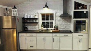 Eine moderne Küche mit weißen Schränken, einem Edelstahl-Kühlschrank, einer Mikrowelle und einem Backofen. Ein Fenster bietet Blick ins Freie, darüber eine Pendelleuchte über der Spüle.