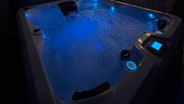 Ein Whirlpool mit blauer Beleuchtung, ein Fernsehbildschirm, der eine Werbeanzeige für eine Uhr zeigt, und ein überdachter Außenbereich mit Lichterketten.