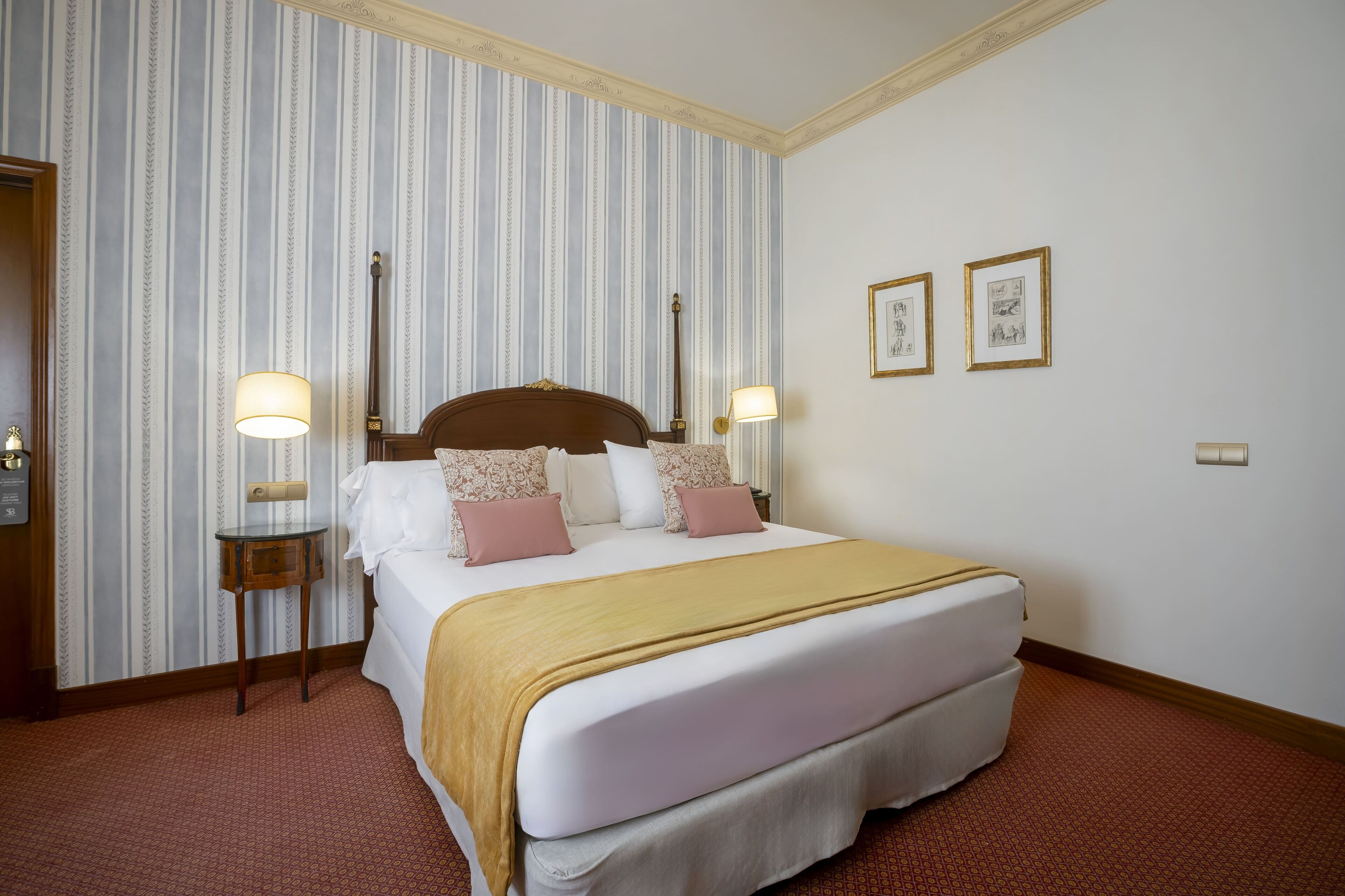 A hotel room with a large bed, two bedside tables with lamps, striped wallpaper, and two framed pictures on the wall.