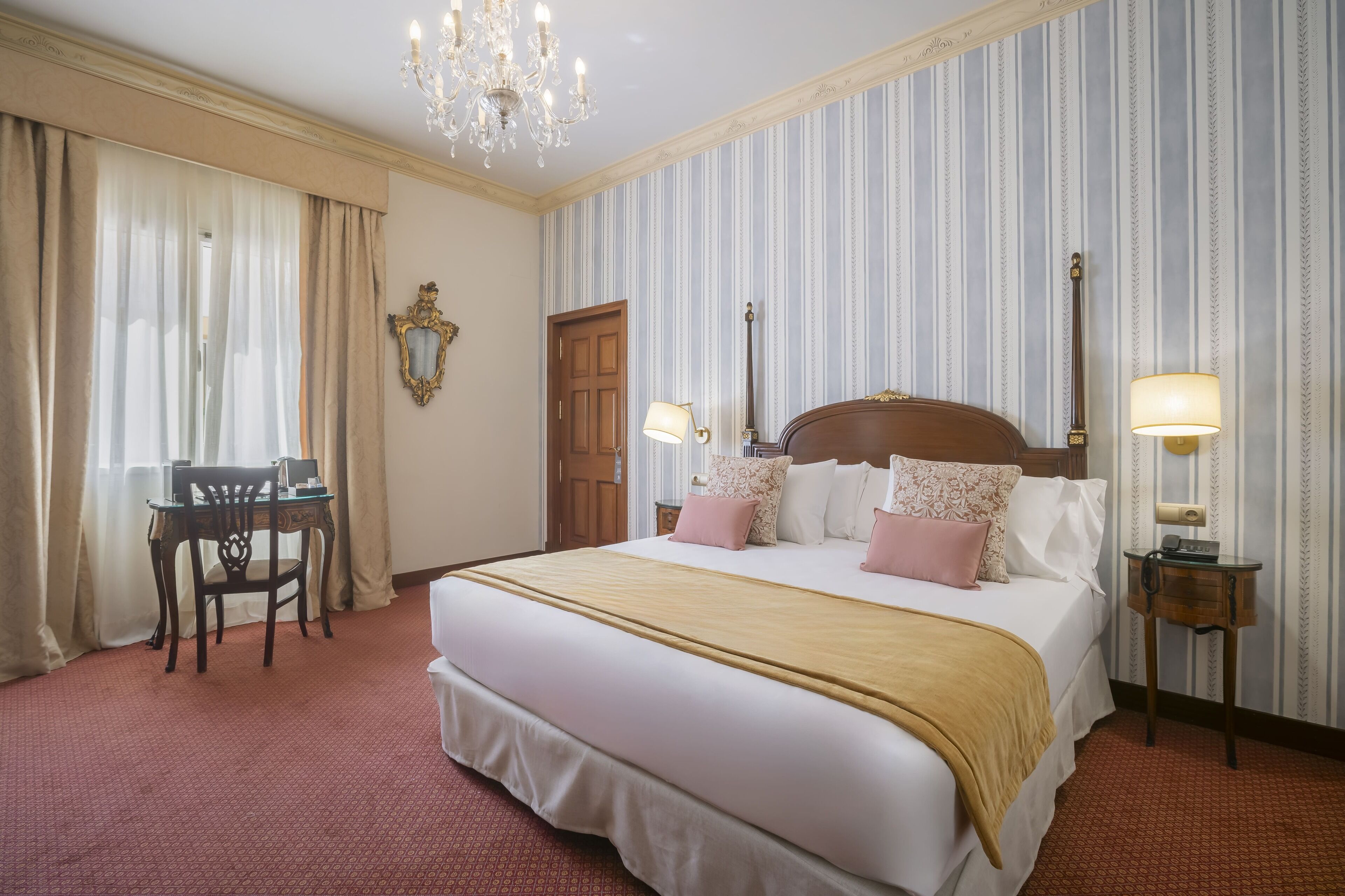 A hotel room with a large bed, two bedside tables with lamps, a wooden chair, a desk, and a chandelier.