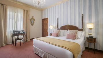A hotel room with a large bed, two bedside tables with lamps, a wooden chair, a desk, and a chandelier.