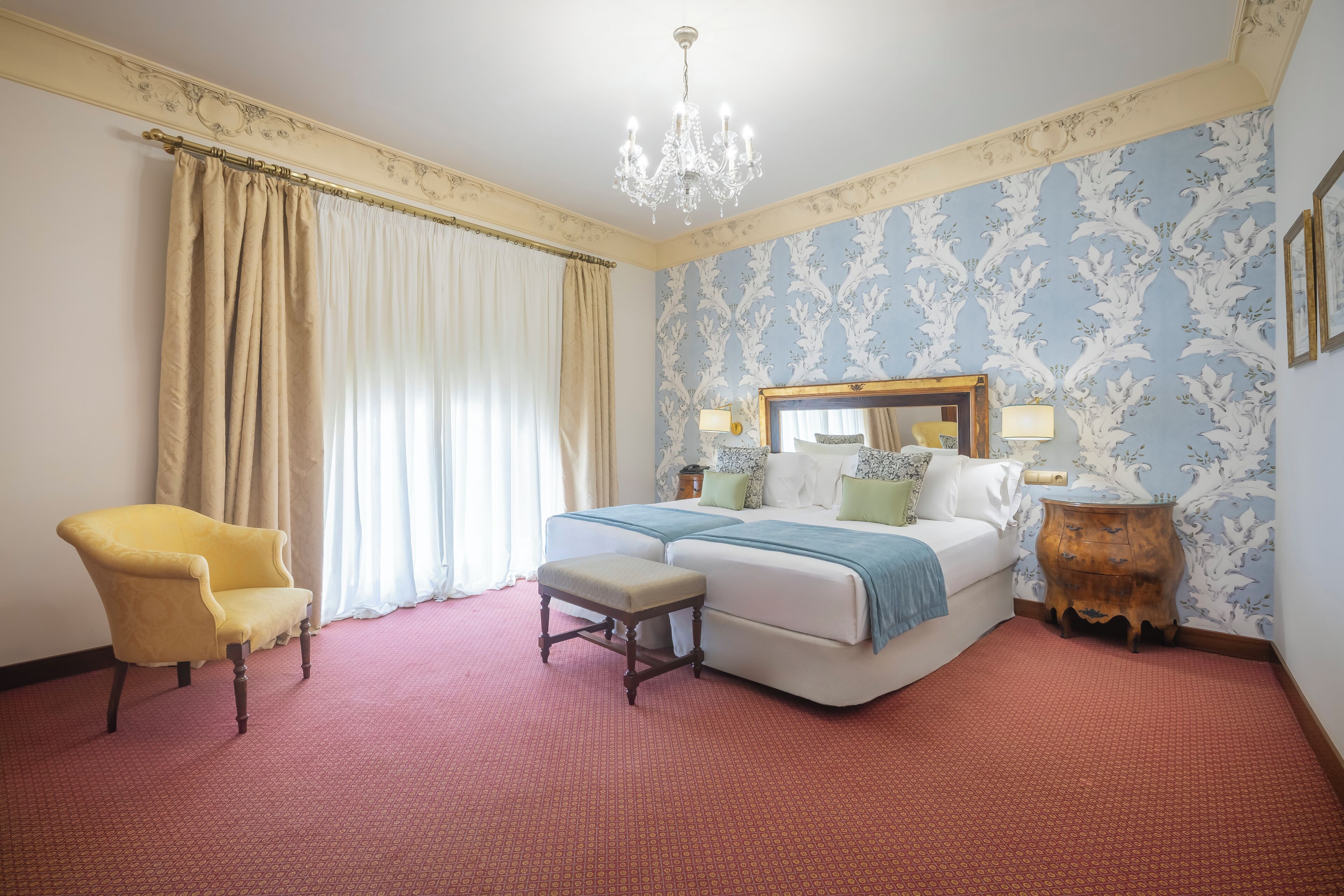 A bedroom with a bed, a chair, a bench, a dresser, and a chandelier.