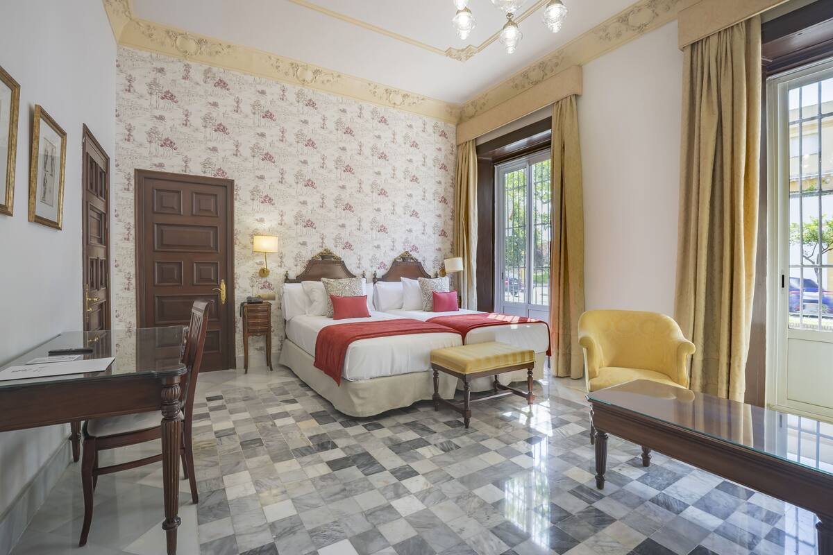 Une chambre avec un sol à carreaux, un lit avec une literie rouge, un fauteuil jaune et un bureau avec une chaise.