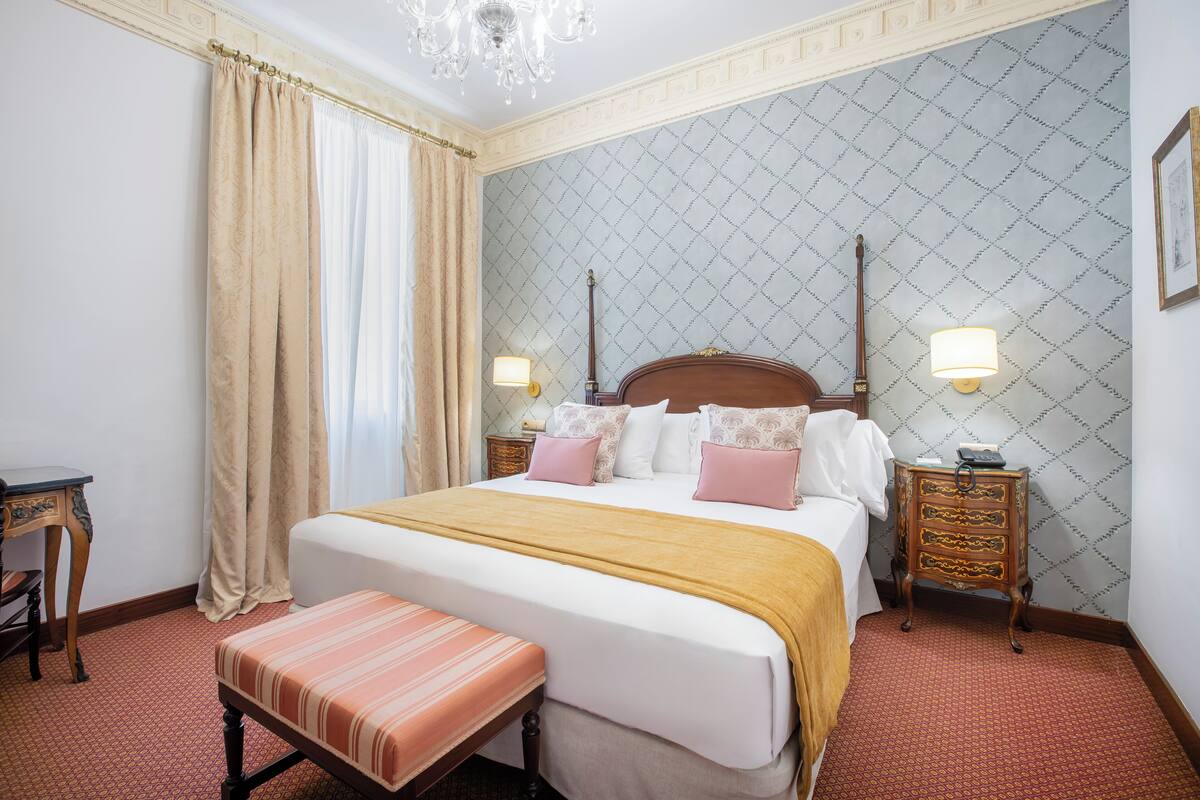 Une chambre d’hôtel avec un grand lit, deux tables de chevet avec des lampes, un banc et un lustre.