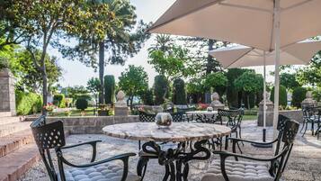 Un patio al aire libre con mesas y sillas redondas, rodeado de follaje y estatuas.