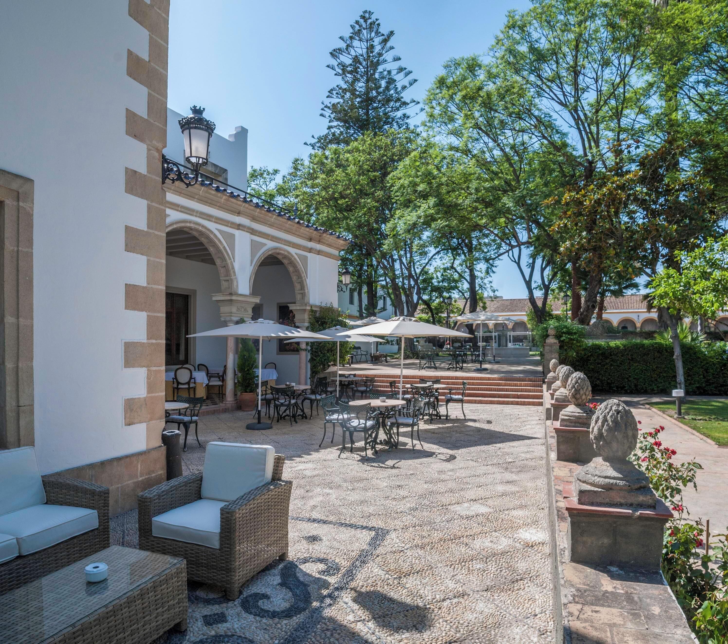Un patio al aire libre con mobiliario de mimbre y sombrillas, rodeado de árboles y un edificio con arcos.