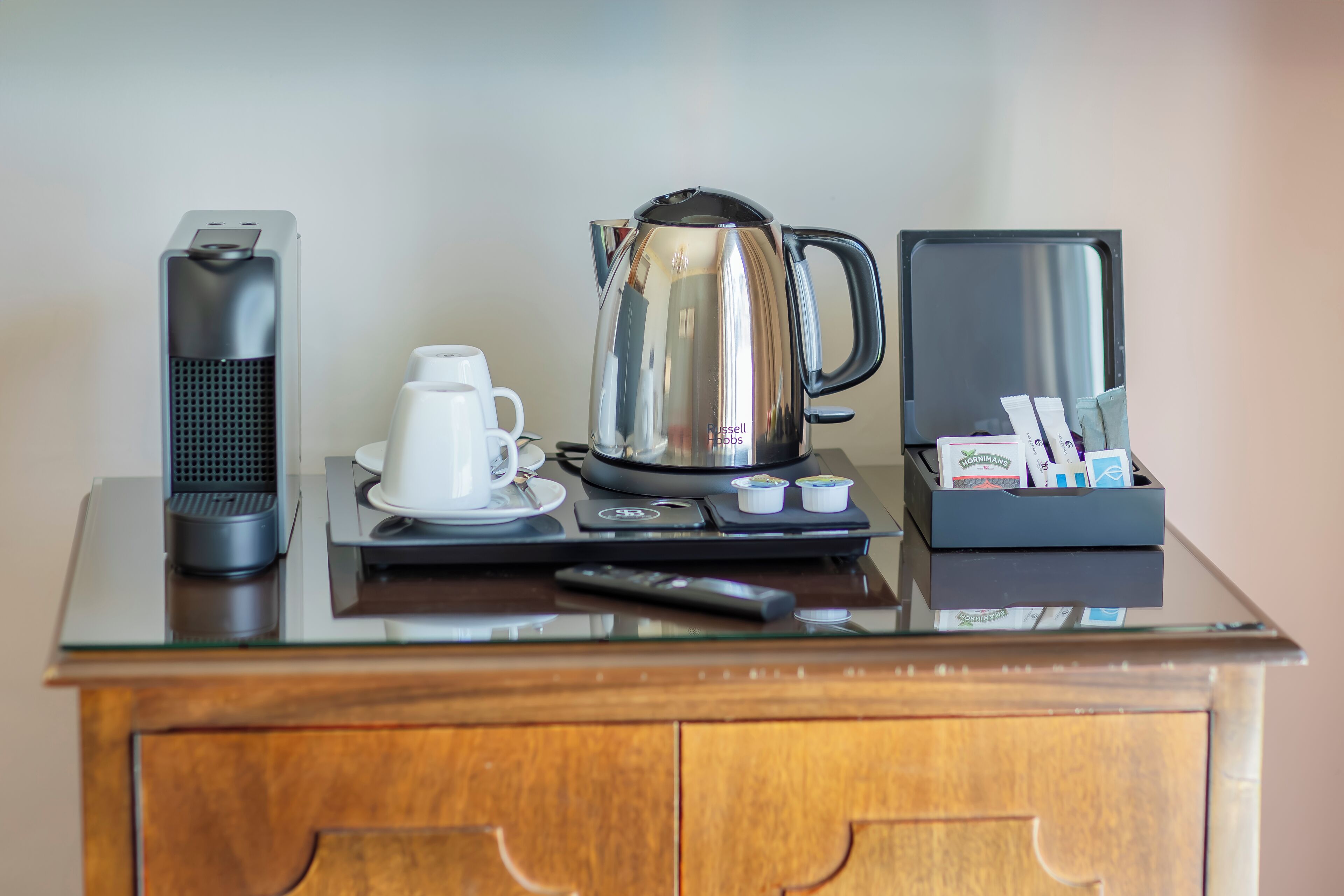 Un coin café comprenant une machine à café, une bouilloire, une tasse et une boîte de capsules de café, le tout posé sur une table à plateau en verre.