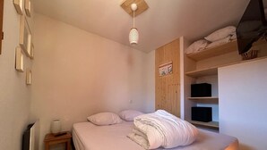 A bedroom with a bed, a bedside table, a radiator, a shelf with boxes, and a poster on the wall. - GREEN THISTLE (Valmeinier)