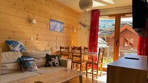 A cozy wooden interior with a sofa, a dining table, and chairs. A painting is on the wall, and there's a view of a wooden building outside. - GREEN THISTLE (Valmeinier)
