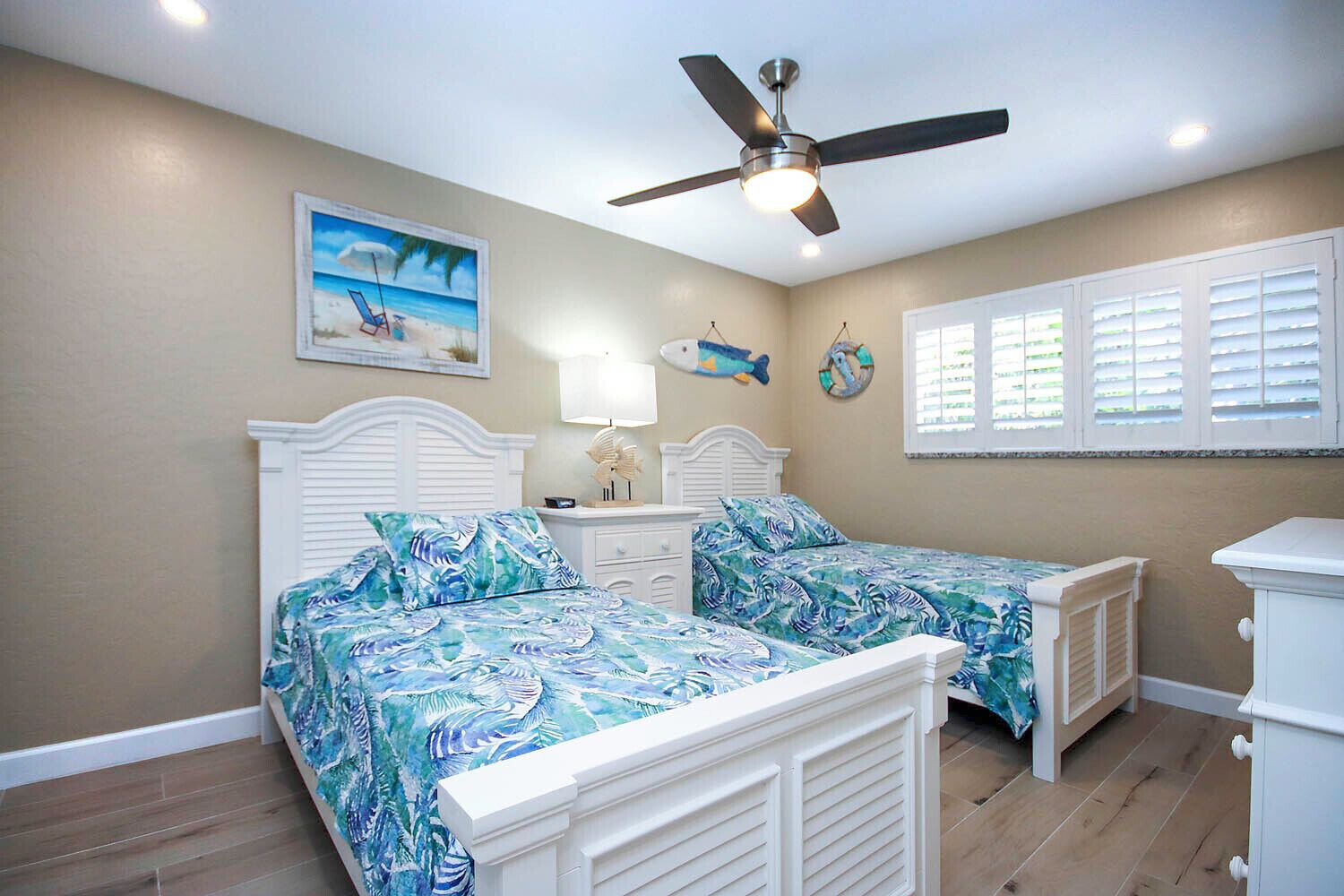 Une chambre avec deux lits, un ventilateur de plafond, un tableau représentant une scène de plage et un objet décoratif en forme de poisson.
