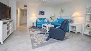 A living room with a blue sofa, a black armchair, a coffee table, and a TV on a stand. There is a ceiling fan, a lamp, and a shelf with decorative items. - Lush 2BR/2BA Sanibel Arms West Garden Unit A4 | Peaceful Oasis Near the Gulf (Sanibel)