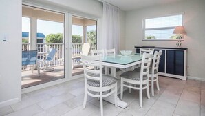 A dining area with a glass table and white chairs, a sliding glass door leading to a balcony, and a window with blinds. - Lush 2BR/2BA Sanibel Arms West Garden Unit A4 | Peaceful Oasis Near the Gulf (Sanibel)