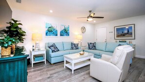 A living room with a light blue sofa, white armchair, and a coffee table. There are two wall-mounted lamps, a ceiling fan, and a clock on the wall. - Cheerful Unit B1 Sanibel Arms West | Just a 2 Minute Walk to the Beach (Sanibel)