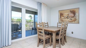 A dining area with a table and chairs, a balcony with a view, and a painting on the wall. - Vibrant Unit B7 · Partial Gulf View | Just a 2 Minute Walk to the Beach (Sanibel)