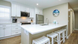 Una cocina moderna con armarios blancos, un refrigerador de acero inoxidable, microondas y cafetera. Hay una isla grande con taburetes de bar y un reloj en la pared.