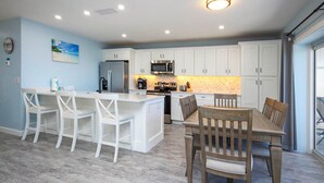 Una cocina moderna con armarios blancos, una isla central con taburetes de bar, una mesa de comedor con sillas y vistas al exterior por una puerta corredera.