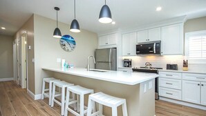 A modern kitchen with a central island, white bar stools, pendant lighting, and a clock on the wall. - Breezy Unit D4 · Sanibel Arms West | Gulffront Breathtaking Gulf Views (Sanibel)