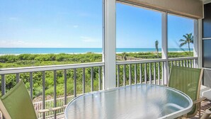 Un balcon avec une table et des chaises en verre, donnant sur une plage bordée de palmiers et sur l’océan.