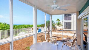 A covered outdoor area with a table and chairs, a ceiling fan, and a view of the ocean and greenery. - Mesmerizing Unit E1 · Sanibel Arms West | Gulffront Magnificent Gulf Views (Sanibel)