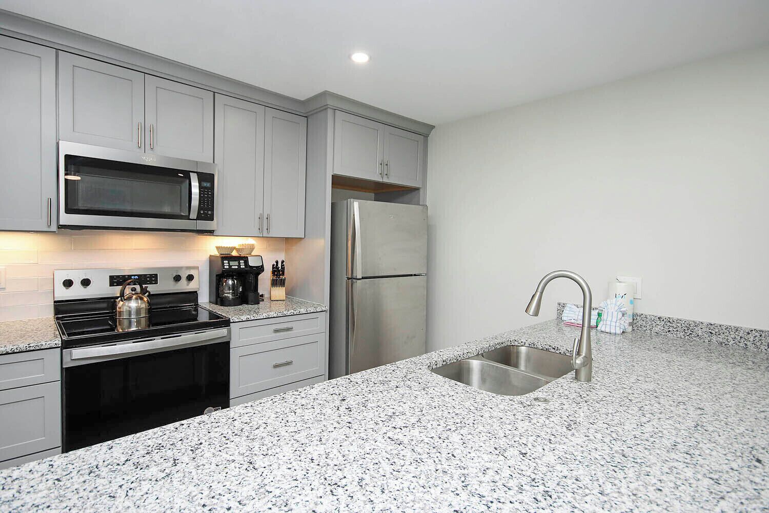 A modern kitchen with grey cabinets, a stainless steel refrigerator, a built-in microwave, a gas stove, and a granite countertop.