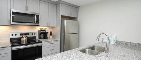A modern kitchen with grey cabinets, a stainless steel refrigerator, a built-in microwave, a gas stove, and a granite countertop.