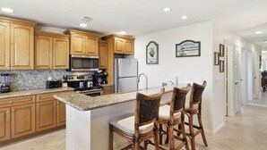 A kitchen with wooden cabinets, granite countertops, a stainless steel refrigerator, and a bar area with wicker chairs. - Breathtaking Unit E7 · Sanibel Arms West | Gulffront Stunning Gulf Views (Sanibel)