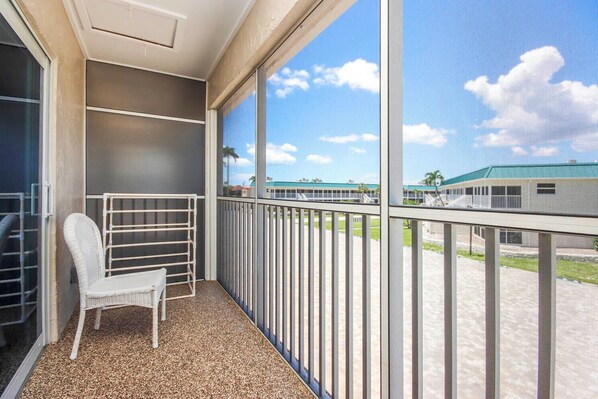 A balcony with a white wicker chair and a drying rack, overlooking a building with a green roof and palm trees.