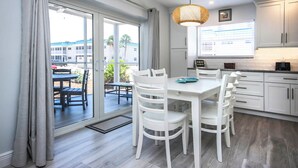 A modern kitchen with white cabinets, a dining area with white chairs, and a large window overlooking a patio. - Breezy Unit G1 · Sanibel Arms West | 2 Minute Stroll to the Gulf (Sanibel)